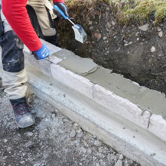 Steinkleber wird mit einer Glättekelle gleichmäßig auf eine Mauer aus Betonsteinen aufgetragen.