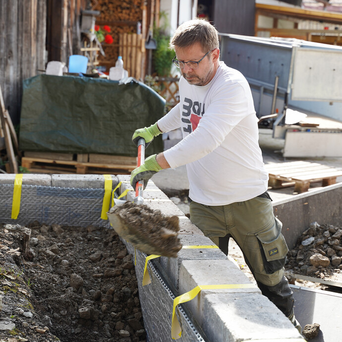 Heimwerker füllt mit Hilfe einer Schaufel die Baugrube an der Gartenmauer mit Aushubmaterial auf.