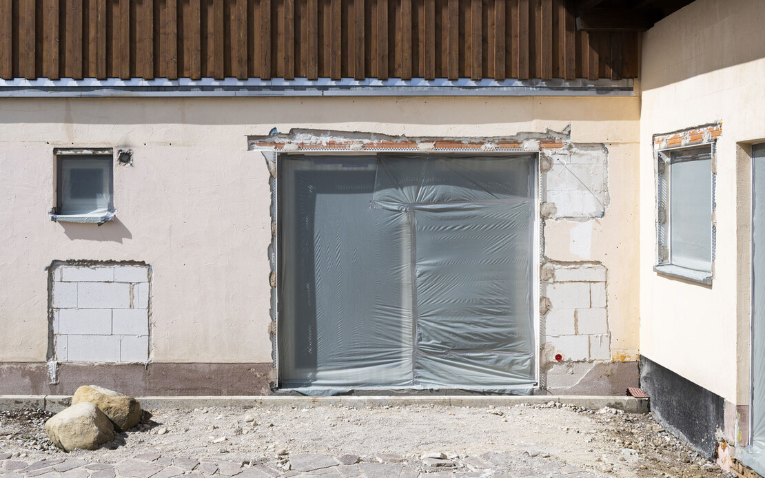 Zeit f&uuml;r eine Sanierung: Alte Hausfassade, die saniert werden muss, mit abgeschlagenem Putz, kleinem Fenster und gro&szlig;er Terrassent&uuml;re.
