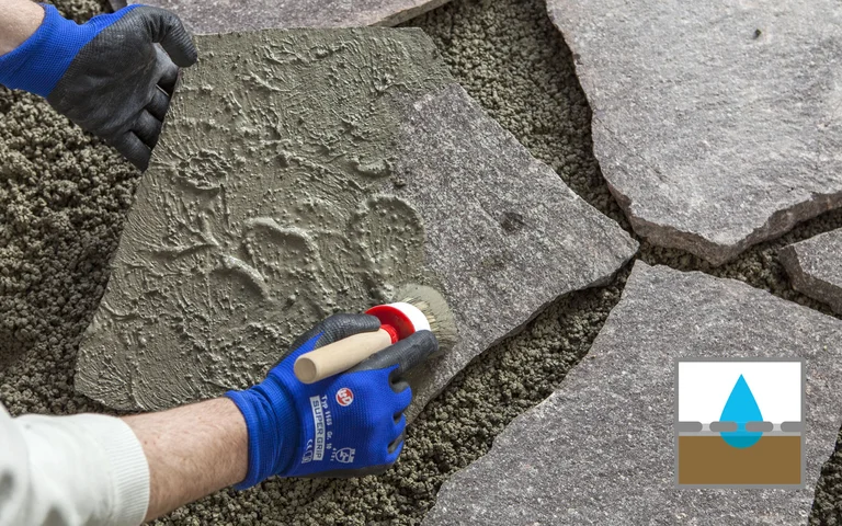 Heimwerker verlegt Natursteinplatten auf Splittuntergrund mit Handschuhen und Pinsel aufgetragenem Mörtel.