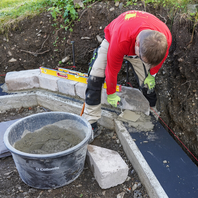 Heimwerker trägt mit einer Kelle Mörtel auf das Fundament auf, um die erste Reihe Steine für die Gartenmauer zu setzen. Daneben steht ein Mörtelkübel mit angerührtem Frischmörtel und ein Mauerstein. Im Hintergrund ist auf der Steinreihe eine Wasserwaage.