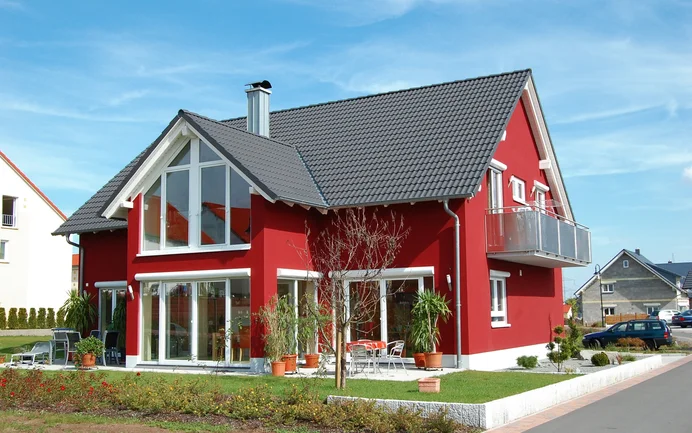 Maison individuelle moderne avec une façade rouge, de grandes fenêtres et un jardin bien entretenu par temps ensoleillé.