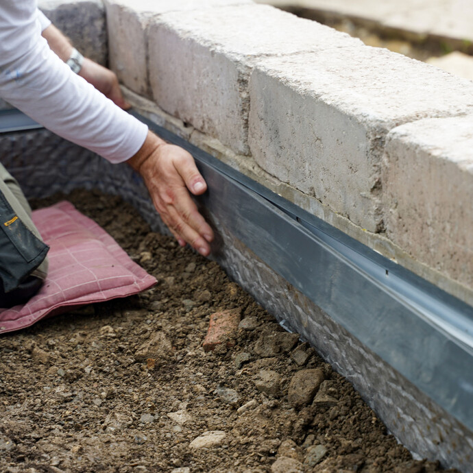 Der Handwerker drückt das Abdeckprofil in das Abschlussprofil ein, damit das Profil fest an der Gartenmauer sitzt. Der Handwerker kniet auf einem rotfarbenem Kissen.