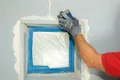 Fensterrahmen wird mit Spachtel und Malerkrepp abgeklebt, um Wandarbeiten vorzubereiten.
