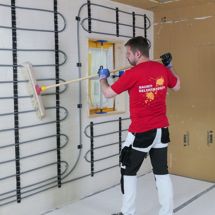 Heimwerker kehrt mit einem Besen eine Wand mit sichtbarer Wandheizung gründlich ab, um sie für den Putz vorzubereiten.