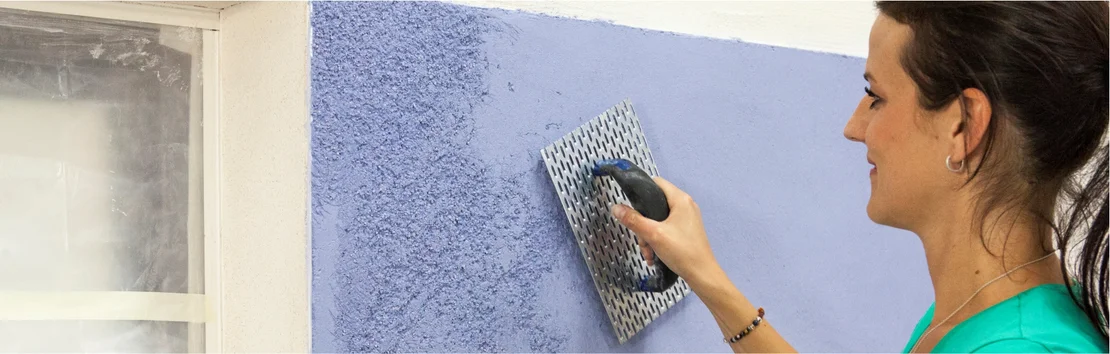Une bricoleuse applique un enduit bleu &agrave; l'aide d'une taloche sur un mur, pr&egrave;s d'une fen&ecirc;tre.