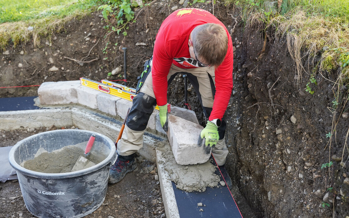 Eine Person setzt einen Mauerstein in einen frischen M&ouml;rtel auf das Fundament im Garten. Eine Wasserwaage und ein M&ouml;rtelk&uuml;bel mit anger&uuml;hrtem Universalm&ouml;rtel und Kelle liegen daneben.