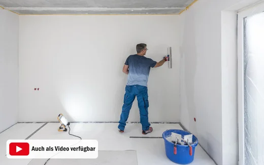 Heimwerker zieht den weißen, feinen Putz eben mit einem Flächenspachtel ab. Um Unebenheiten zu sehen, wird die Wand von einem Baustrahler angestrahlt. Diese Anleitung für Heimwerker gibt es mit Text und Bild und ist auch als Video verfügbar.