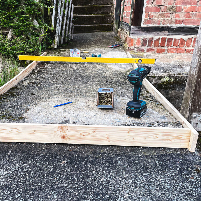 Holzschalung aus Kanthölzern wird auf dem Untergrund ausgerichtet und mit Wasserwaage kontrolliert, Akkuschrauber und Schrauben liegen bereit.