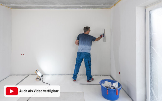 Heimwerker zieht den weißen, feinen Putz eben mit einem Flächenspachtel ab. Um Unebenheiten zu sehen, wird die Wand von einem Baustrahler angestrahlt. Diese Anleitung für Heimwerker gibt es mit Text und Bild und ist auch als Video verfügbar.