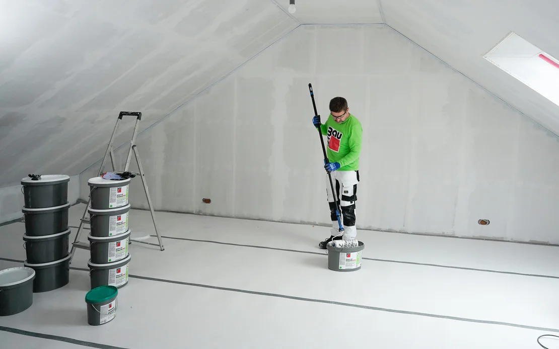 Heimwerker tr&auml;gt wei&szlig;en Innenanstrich mit Farbrolle in einem fertig verputzten Dachzimmer auf.