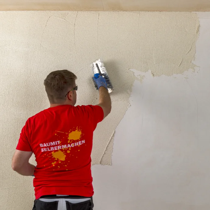 Mann trägt Putz mit Kelle auf Wand auf, Baumit-Logo auf T-Shirt hinten sichtbar, Innenrenovierung.