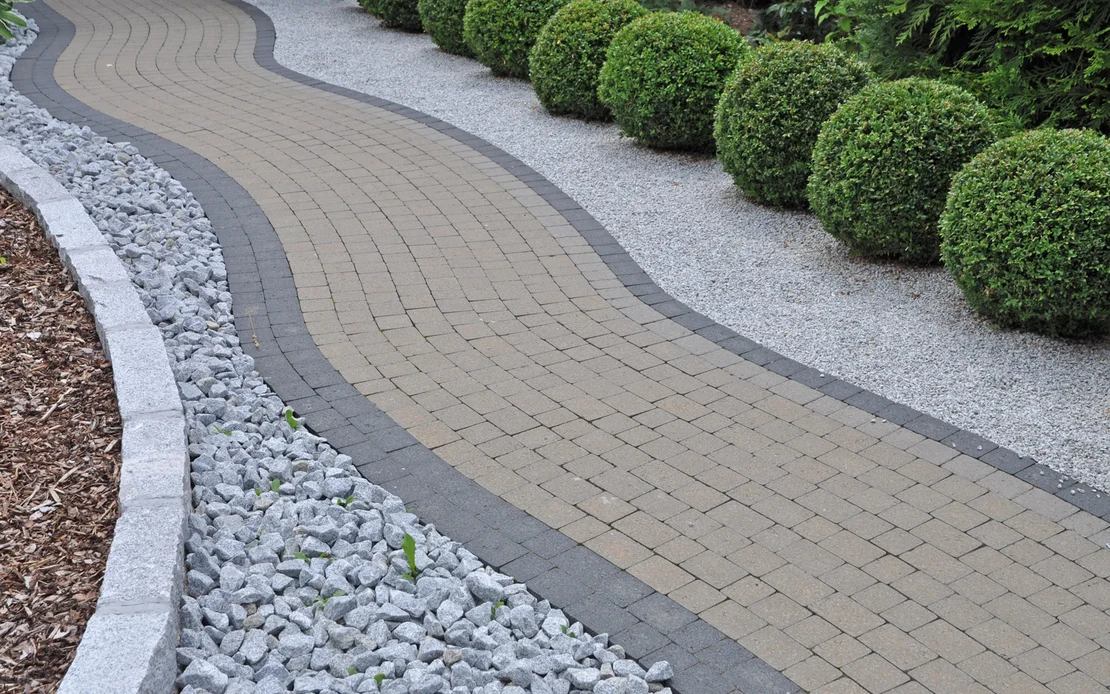 Gepflasterter Gartenweg mit dekorativen Randsteinen und Kiesbeeten, ideal zur Versch&ouml;nerung von Au&szlig;enanlagen.