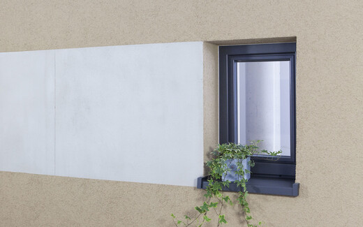 Fassade mit beigem Oberputz und verputzter Fläche in Betonoptik neben einem Fenster mit Blumenschmuck.